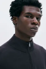 Buzo de punto fino color marrón oscuro, con cuello alto acanalado y medio cierre frontal. Presenta remates acanalados en puños y bajo.