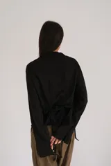 Camisa negra de lino con cuello camisero, manga larga y cierre frontal con botones de nácar. Presenta un diseño con cordones laterales que permiten fruncir la prenda para un ajuste personalizado.