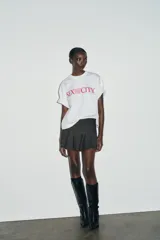 Camiseta blanca de manga corta y cuello redondo, con corte regular y bajo recto. Presenta el logo de "Sex & The City" estampado en color fucsia en el frente.