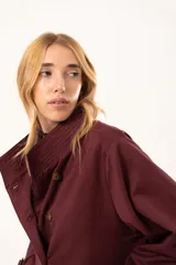 Trench de corte holgado color bordó, con cuello camisero, cierre frontal con botones y cinturón ajustable con hebilla. Presenta mangas largas con detalles de correas en los puños y bolsillos laterales.