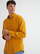 Camisa de manga larga color mostaza confeccionada en pana fina, con bolsillo en el pecho, botones de pasta, bordado en el ruedo y etiqueta de marca en la tapeta de los botones.