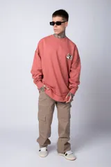 Buzo de tejido friza color crema, de corte oversize y cuello redondo. Presenta un pequeño bordado o parche en el pecho izquierdo con un diseño de cabeza de animal en tonos rojizos/terracota.