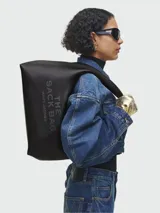 Bolso de hombro Marc Jacobs modelo The Sack, confeccionado en cuero negro con textura granulada. Presenta el nombre del modelo y la marca grabados en relieve en el frente. Diseño minimalista y espacioso con correa integrada al cuerpo del bolso.