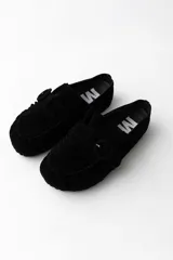 Slipper tipo zueco de gamuza negra, con diseño de puntera redondeada y detalle de hebilla forrada en el empeine. Presenta costuras decorativas en el borde y suela plana.