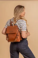 Mochila tipo cartera con tapa, confeccionada en símil cuero color suela. Cuenta con doble compartimento, división interna, cierre de seguridad y bolsillo trasero con cierre. Incluye una correa frontal decorativa con tachas metálicas.