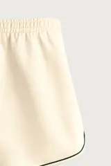 Bermuda de corte relajado confeccionada en tejido de algodón, color beige con vivos en contraste color verde oscuro. Cuenta con cintura elástica ajustable con cordón, bolsillos frontales y bolsillos traseros de plastrón.
