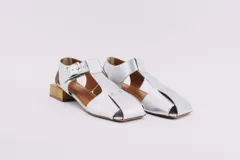 Sandalias de cuero color plateado con diseño de punta cerrada y talón descubierto. Presentan una tira con hebilla ajustable sobre el empeine y un taco cuadrado bajo con acabado de madera.