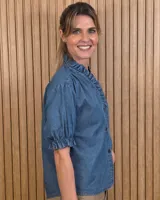 Camisa de denim de algodón celeste, con cierre frontal de botones, cuello alto con volados y mangas cortas abullonadas con puños elastizados fruncidos.