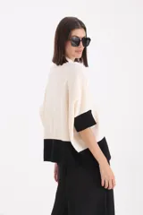 Sweater de punto bicolor con cuello alto, mangas cortas y corte holgado. Presenta un diseño con bloque de color negro en el ruedo y las mangas, y un detalle decorativo de cinto de cuero sintético con hebilla en el frente.