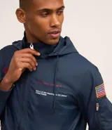 Campera rompevientos azul marino con capucha, cierre frontal y bolsillos laterales. Presenta estampado de NASA en el pecho, parche de bandera estadounidense en la manga y bolsillo utilitario en el brazo.