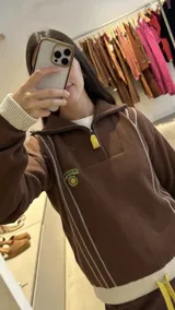 Buzo de punto color marrón con cuello polo y cierre frontal parcial. Presenta detalles de líneas verticales en color crema, puños y ruedo acanalados en tono crema, y un pequeño parche bordado en el pecho. Incluye tirador de cierre en color amarillo.