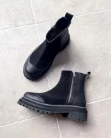 Botas cortas de cuero vacuno color negro, con diseño combinado de gamuza y cuero texturizado con efecto reptil en el talón. Presentan cierre lateral metálico, puntera reforzada y suela track dentada con costura visible.