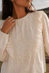 Blusa de color beige con bordados florales en el frente, escote redondo y mangas largas con puños elastizados. Presenta volados decorativos a los lados del pecho.