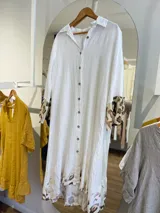 Vestido camisero largo de lino blanco con botones, cuello clásico y mangas largas con puños estampados con animal print. Tiene un corte irregular con el frente más corto que la parte trasera y estampado animal print en el ruedo.