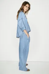 Camisa de denim en tono azul claro, con cuello solapa, escote en pico y cierre frontal con botones. Presenta mangas anchas por debajo del codo y un diseño de bajo asimétrico.