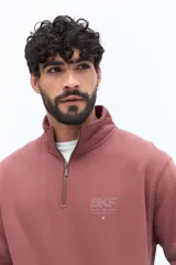 Buzo de algodón color terracota, con cuello alto y cierre parcial frontal. Presenta puños y cintura ajustados, bolsillos laterales y un pequeño logo bordado en el pecho.