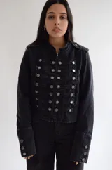 Chaqueta de estilo militar confeccionada en denim negro con efecto lavado. Presenta un diseño estructurado con cierre frontal de cremallera, cuello mao y múltiples botones metálicos decorativos dispuestos en filas horizontales. Incluye detalles de charreteras en los hombros y puños con botones.