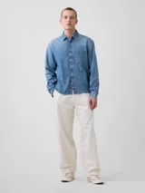 Camisa de jean de corte recto y calce relajado, con cierre frontal de botones metálicos, cuello clásico y un bolsillo frontal en el pecho.