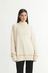 Buzo color crema de punto con cuello alto, costuras a mano en color naranja y bordado de marca en la manga. Corte oversize.