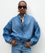Campera bomber de jean azul con cuello redondo, mangas abullonadas con puños elásticos, cintura ajustable y cierre frontal con botones a presión.