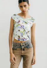 Top corto de jersey con estampado abstracto floral en tonos pastel (blanco, lila, verde, marrón). Presenta un diseño cruzado asimétrico en el busto que crea un escote tipo *off-shoulder* en un lado y manga corta en el otro.