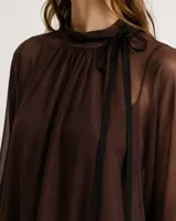 Camisa de gasa color marrón oscuro, de corte oversize y cuello alto fruncido con cordones colgantes. Presenta mangas largas abullonadas con puños ajustados por lazos y dobladillo asimétrico con terminación en puntas.