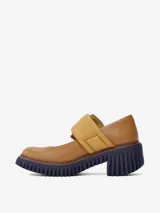 Zapato tipo Mary Jane de cuero color camel, con suela gruesa y dentada color azul marino. Presenta una tira ancha con hebilla metálica dorada y un detalle de ribete azul claro en el lateral de la tira.