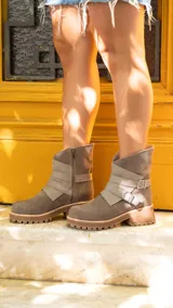 Botas cortas de estilo urbano, confeccionadas en una combinación de gamuza y cuero vacuno en tonos tierra. Presentan un diseño con tiras decorativas cruzadas en el empeine, hebillas metálicas ajustables, cierre lateral y suela de goma con plataforma dentada.