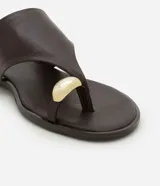 Sandalia plana Satinato confeccionada en cuero color marrón, con diseño de tira para el dedo y detalle metálico dorado redondo. Cuenta con suela baja.