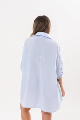 Vestido camisero de lino color celeste, con escote en V, cuello clásico, cierre frontal de botones y mangas tres cuartos regulables. Diseño holgado con corte asimétrico.