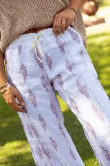 Pantalón de lino de corte amplio y tiro medio, con cintura elástica y cordón ajustable. Presenta un estampado lineal con motivos decorativos en tonos violetas sobre fondo blanco.