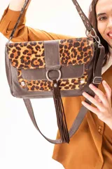 Cartera de cuero vacuno color marrón chocolate combinada con pelo vacuno en estampa leopardo. Presenta una tapa frontal con apliques metálicos, un fleco central decorativo y cierre principal. Incluye una correa corta con tachas y una correa larga extensible y desmontable.