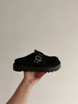 Sueco de plataforma color negro, confeccionado en material sintético tipo gamuza. Presenta una tira frontal con hebilla metálica y ojales decorativos, con suela dentada de goma.