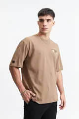 Remera oversize color marrón claro con estampado gráfico en la espalda que incluye frases como "Stay Original Forever", "All About Having Fun" y el logo de Mistral, junto con otros garabatos y stickers.