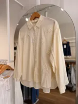Camisa de corte oversize color beige claro, con mangas largas y cuello camisero clásico. Presenta delicados bordados florales en relieve en la parte superior del frente y abotonadura frontal con botones blancos.