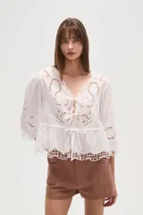 Blusa blanca de algodón con bordados tipo broderie anglaise, escote en V con lazos ajustables en el centro del busto y peplum con terminación festoneada. Mangas tres cuartos.