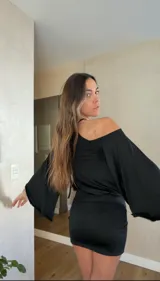 Vestido corto negro con cuello halter, hombros descubiertos y mangas largas acampanadas. Presenta un diseño ajustado en la cintura y falda con frunces laterales.