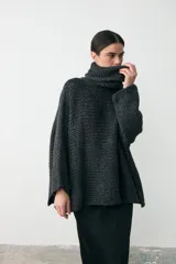 Sweater tipo poncho de tejido grueso en color gris oscuro, con cuello alto tipo polera y mangas anchas. Presenta un diseño holgado y texturizado.