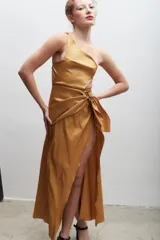 Vestido largo dorado de corte asimétrico con un solo hombro, detalle drapeado en la cintura, lazo lateral y tajo frontal pronunciado.