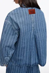 Chaqueta corta de denim en color azul con diseño de rayas verticales blancas, cuello camisero, bolsillos delanteros de parche y cierre frontal con botones metálicos.