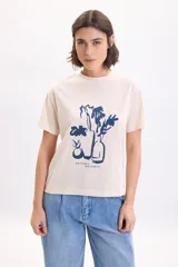 Remera color crudo de manga corta, con cuello redondo y estampa frontal en color azul de botellas y flores junto con la frase "Natural Balance".