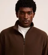 Saco deportivo negro de manga larga, confeccionado en tejido fleece, con cuello alto y cierre frontal parcial de cremallera. Presenta un corte recto y costuras horizontales a la altura del pecho.