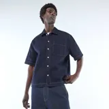 Camisa de jean color azul oscuro con costuras en contraste, cuello clásico, cierre frontal con botones y bolsillo frontal en el pecho.