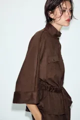 Camisa de efecto ante en color marrón, con cuello subido, mangas tres cuartos con vuelta y cierre frontal con cremallera. Presenta falsos bolsillos con solapa en el pecho y cintura ajustable con cordones del mismo tejido.