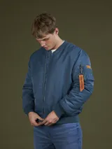 Campera tipo bomber color azul, con cierre frontal, puños y cintura acanalados. Presenta un bolsillo utilitario en la manga con una cinta decorativa naranja con la inscripción Benson & Thomas.