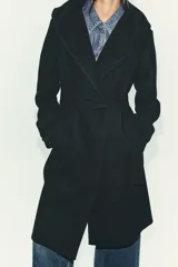 Abrigo de paño negro con capucha, cierre frontal cruzado con botones y cinturón de lazada en el mismo tejido. Presenta manga larga con trabillas en los puños y bolsillos laterales.