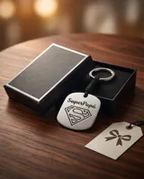 Llavero de acero inoxidable con placa cuadrada grabada con el logo de Superman y la frase Super Papá, unido a una correa trenzada de color negro y argolla metálica circular.