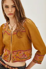 Chaqueta corta color mostaza con mangas abullonadas y bordados étnicos en tonos rosados y rojos en el frente, puños y hombros. Presenta un cierre frontal con detalles de cuentas.