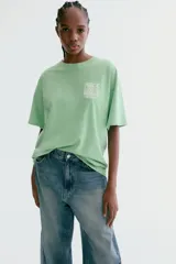 Remera oversized verde de punto de algodón, con hombros caídos, cuello redondo acanalado y estampa pequeña en el pecho con la frase 'Peace Love & Music Woodstock'.