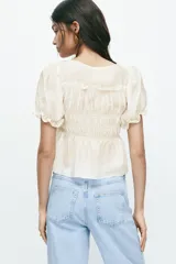 Blusa color beige, de mezcla de lyocell, con escote redondo, mangas cortas abullonadas y busto fruncido con tiras decorativas.